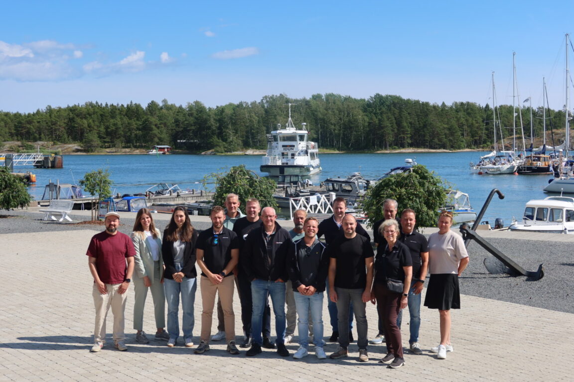 Ryhmäkuva, jossa parikymmentä henkilöä seisoo Kasnäsin satamassa ja katsoo suoraan kameraan. aurinkoisessa säässä.