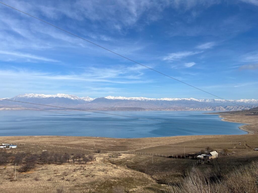 Sininen taivas, horisontti täynnä lumihuippuisia vuoria, vuoriston edessä vesialue ja sen rannoilla harvaa asutusta.